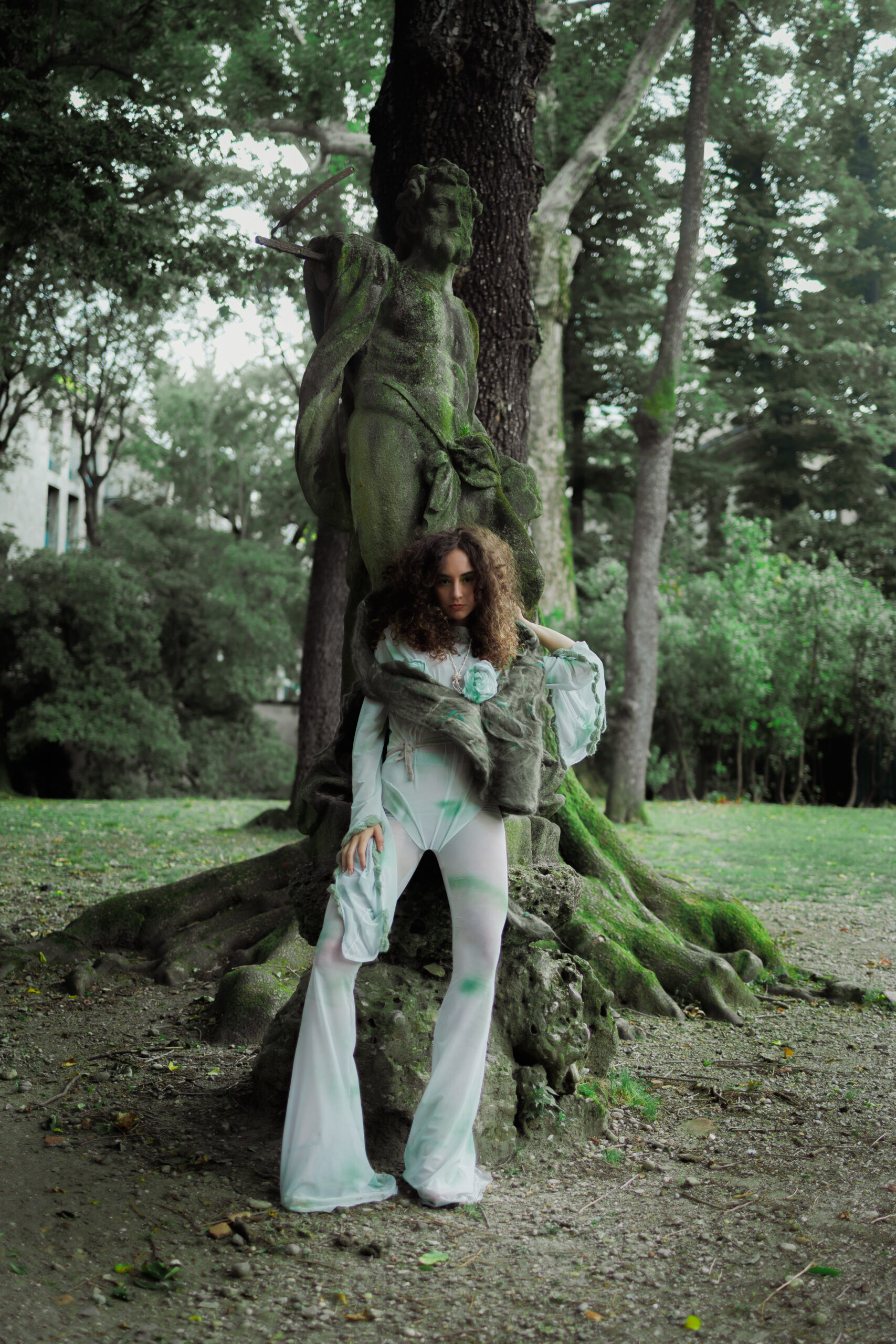 “Model wearing a designer oval-shaped linen gauze cape with wet-felted layers and ribbon closure, styled with a bi-elastic tulle bodice, transparent spray-printed green and grey leggings flared at the hem, and a long-sleeved shirt featuring circular side cuts, crochet leaf details, and crinoline structure.”